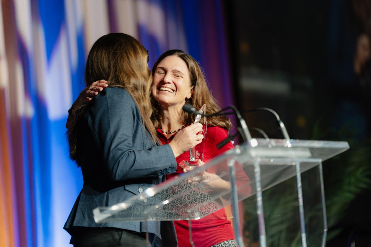 Two individuals conversing near a podium, one holding a microphone, and both dressed in professional attire. Two individuals conversing near a podium, one holding a microphone, and both dressed in professional attire.