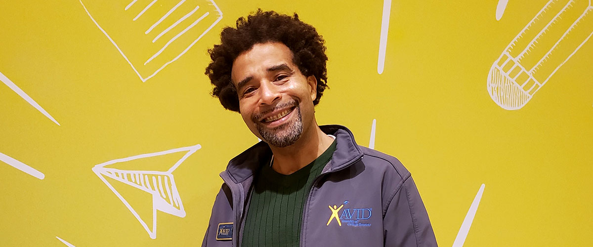 A person wearing a grey jacket with a yellow background displaying an AVID logo and various white line drawings.