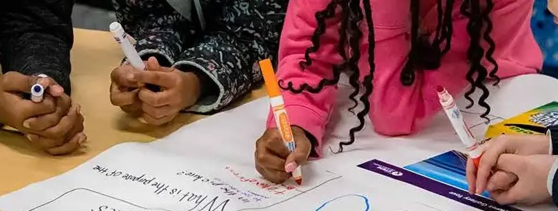 A close-up image of several individuals collaborating on a project at a table, using markers to write on a large sheet of paper. A close-up image of several individuals collaborating on a project at a table, using markers to write on a large sheet of paper.
