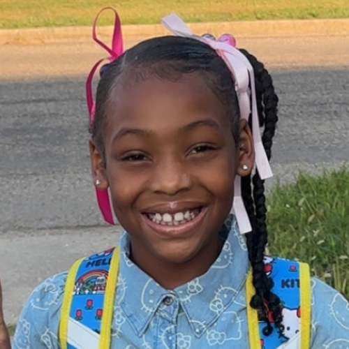 Person with braided hair tied with pink bows, wearing a light blue shirt with patterns and carrying a colorful backpack, standing outdoors.