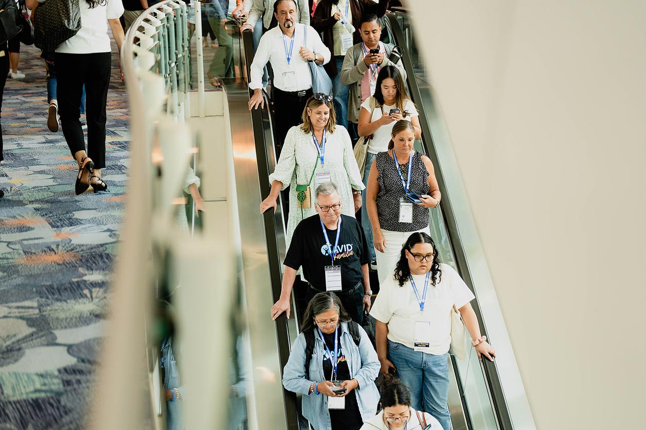 A group of people are descending on an escalator in an indoor setting wearing lanyards and badges, seemingly attending a conference or event. A group of people are descending on an escalator in an indoor setting wearing lanyards and badges, seemingly attending a conference or event.
