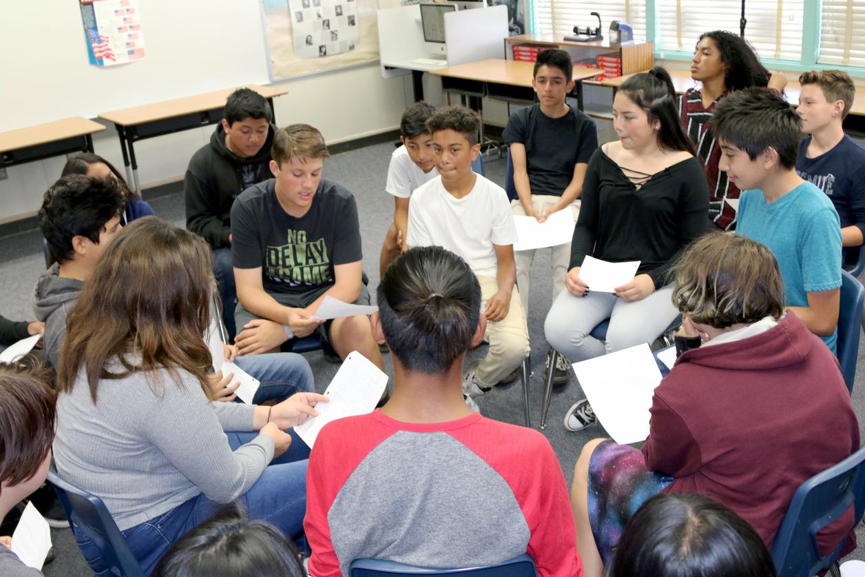 A group of students sitting in a classroom arranged in a circle, holding papers and engaging in discussion. A group of students sitting in a classroom arranged in a circle, holding papers and engaging in discussion.