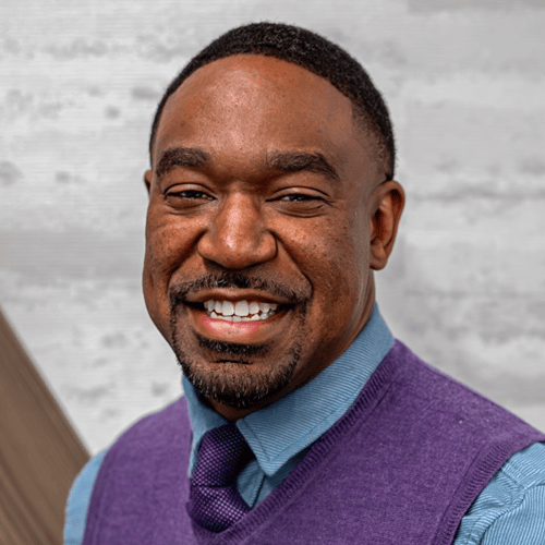 Individual in a blue shirt and purple sweater vest against a light gray background. Individual in a blue shirt and purple sweater vest against a light gray background.