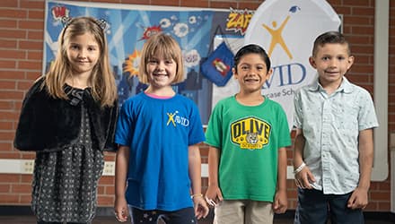 Four children standing in front of a backdrop with school-themed decorations. Four children standing in front of a backdrop with school-themed decorations.