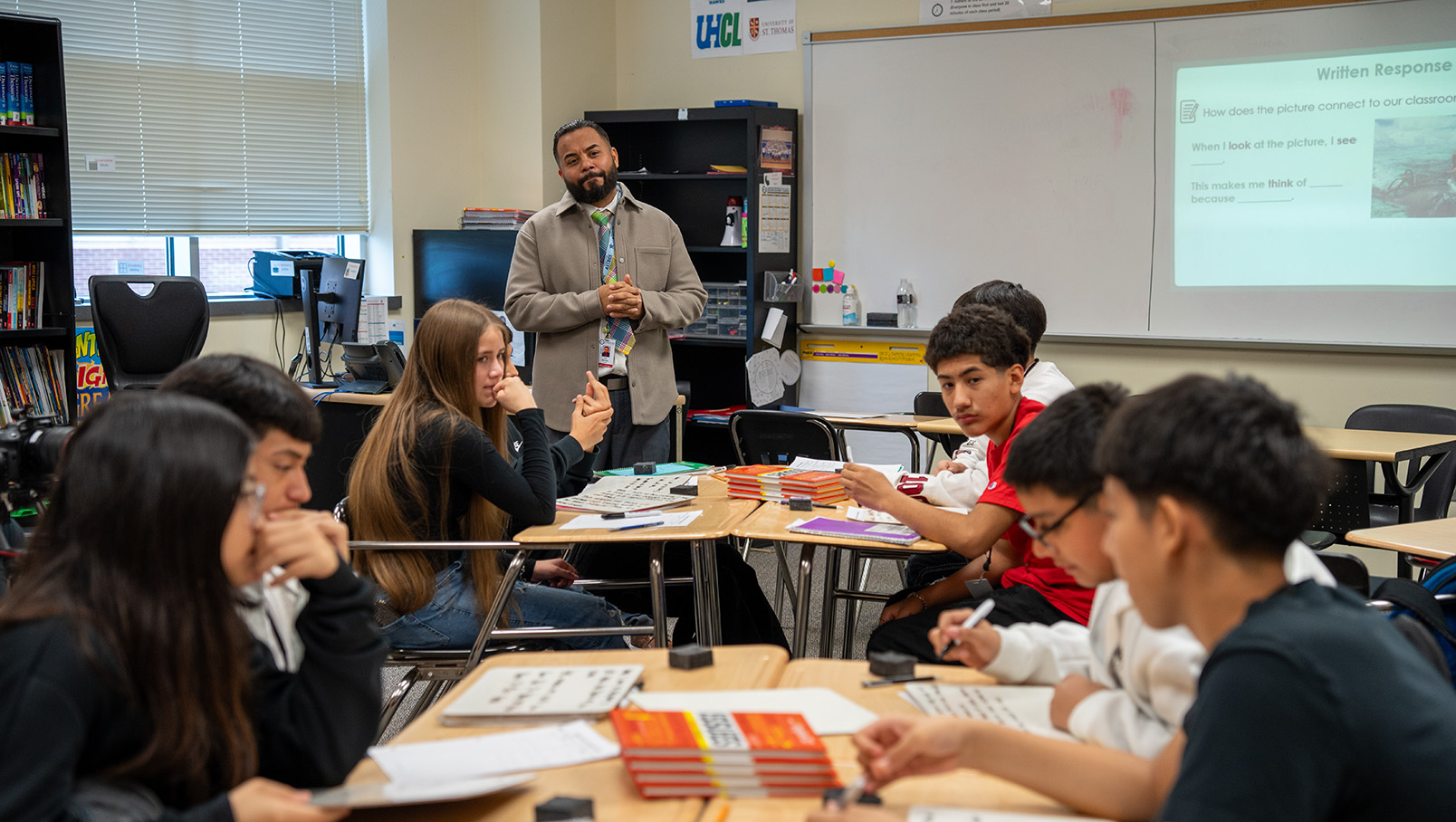A classroom setting with a teacher standing and students seated around a table engaged in discussions with books and papers on the desks.