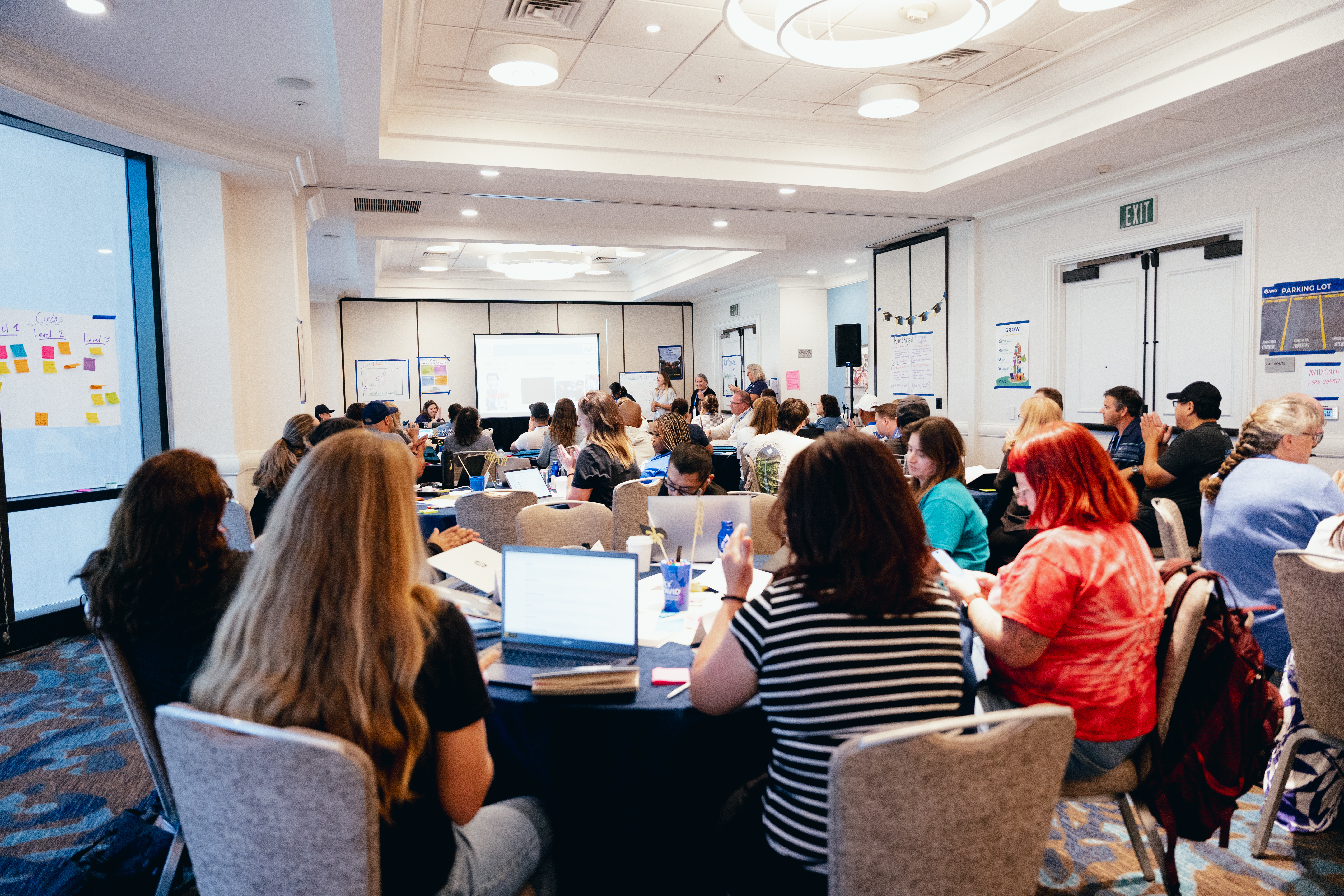 A conference room filled with people seated at tables, attentively watching a presentation on a screen at the front. The room is brightly lit with papers and laptops on the tables.