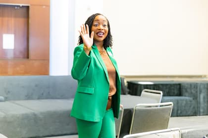 Person wearing a green suit and gesturing with an open hand while standing in a room with grey couches and chairs. Person wearing a green suit and gesturing with an open hand while standing in a room with grey couches and chairs.