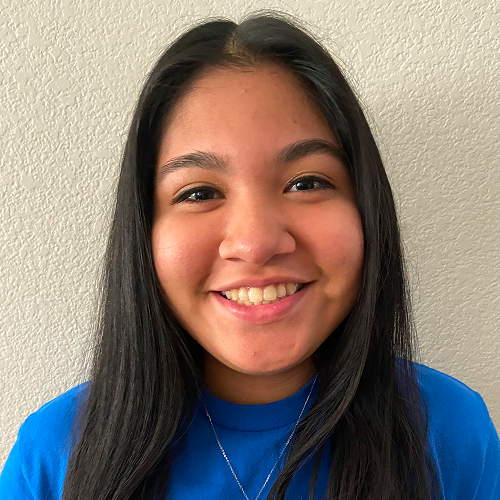 Person with long, dark hair wearing a blue shirt against a light-colored textured wall.
