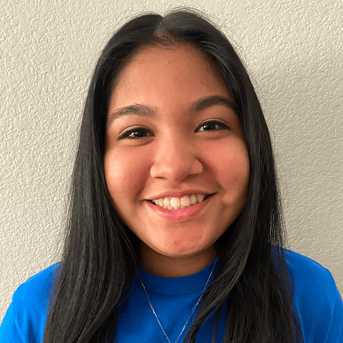 Person with long, dark hair wearing a blue shirt against a light-colored textured wall. Person with long, dark hair wearing a blue shirt against a light-colored textured wall.
