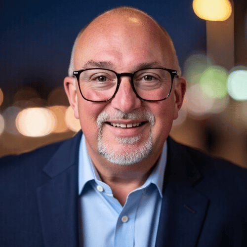 Individual wearing a blue suit jacket over a light blue shirt, with blurred background lights.