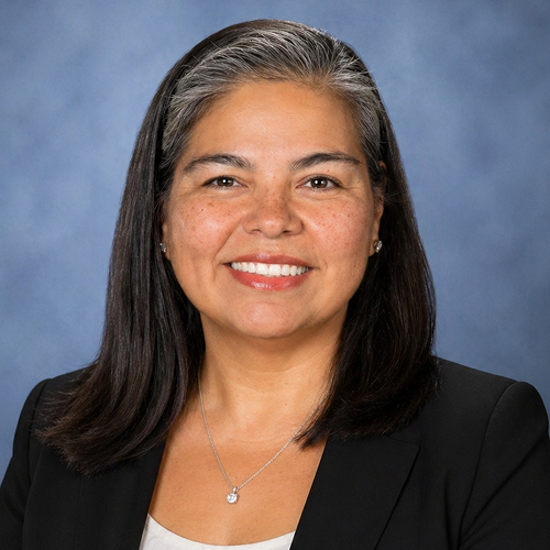 Person with shoulder-length dark hair, wearing a black blazer and a necklace against a neutral background. Face is blurred for privacy.