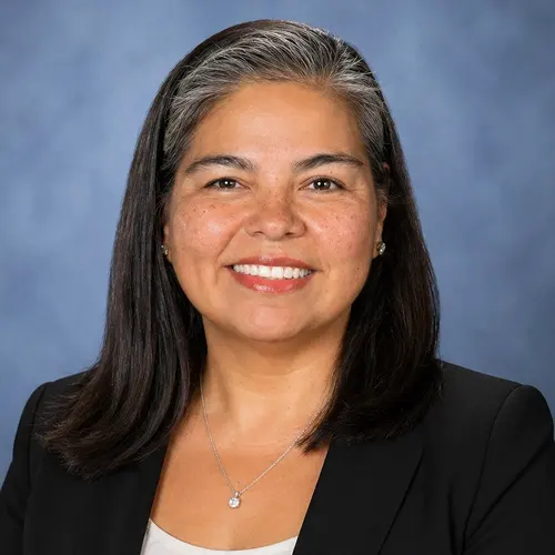 Person with shoulder-length dark hair, wearing a black blazer and a necklace against a neutral background. Face is blurred for privacy. Person with shoulder-length dark hair, wearing a black blazer and a necklace against a neutral background. Face is blurred for privacy.