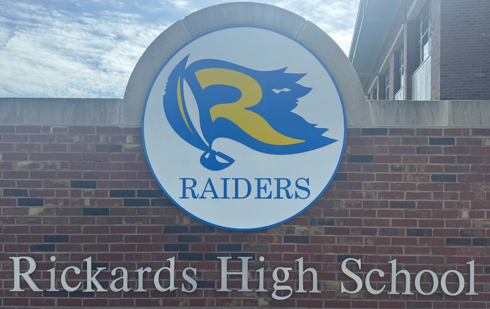 Rickards High School sign with a brick wall and a circular Raiders logo featuring a stylized letter 'R' with blue and yellow colors.