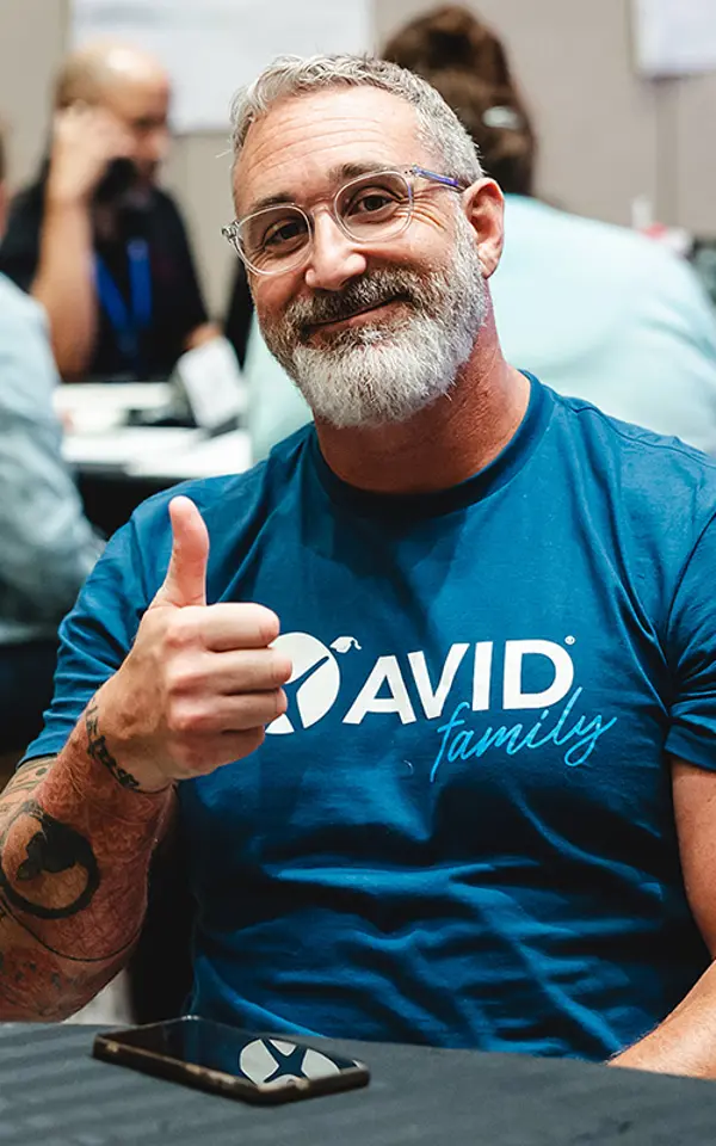 Person in blue AVID family shirt giving a thumbs up, seated at a table with phone.