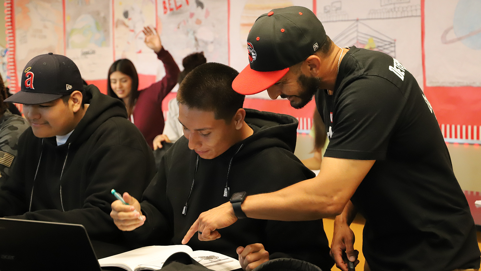 A group of people in a classroom setting, with one person guiding another on their work.