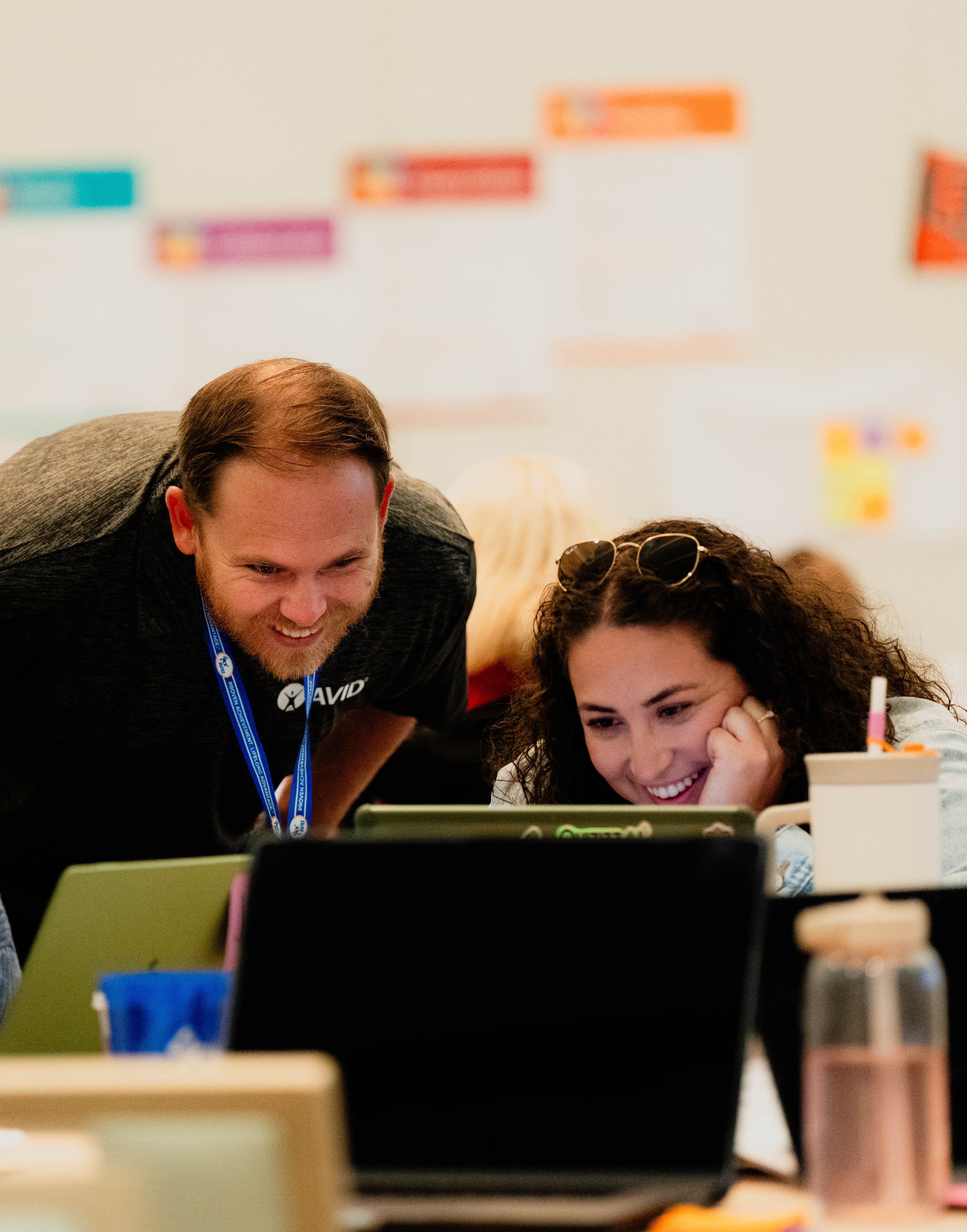 Two individuals interacting at a table with laptops and other items, with faces blurred for privacy. Various posters and signs can be seen on the wall in the background.