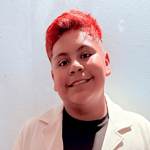 The image features a person with bright red hair wearing a light-colored jacket and a dark shirt, standing against a light gray textured wall. The face is blurred. The image features a person with bright red hair wearing a light-colored jacket and a dark shirt, standing against a light gray textured wall. The face is blurred.