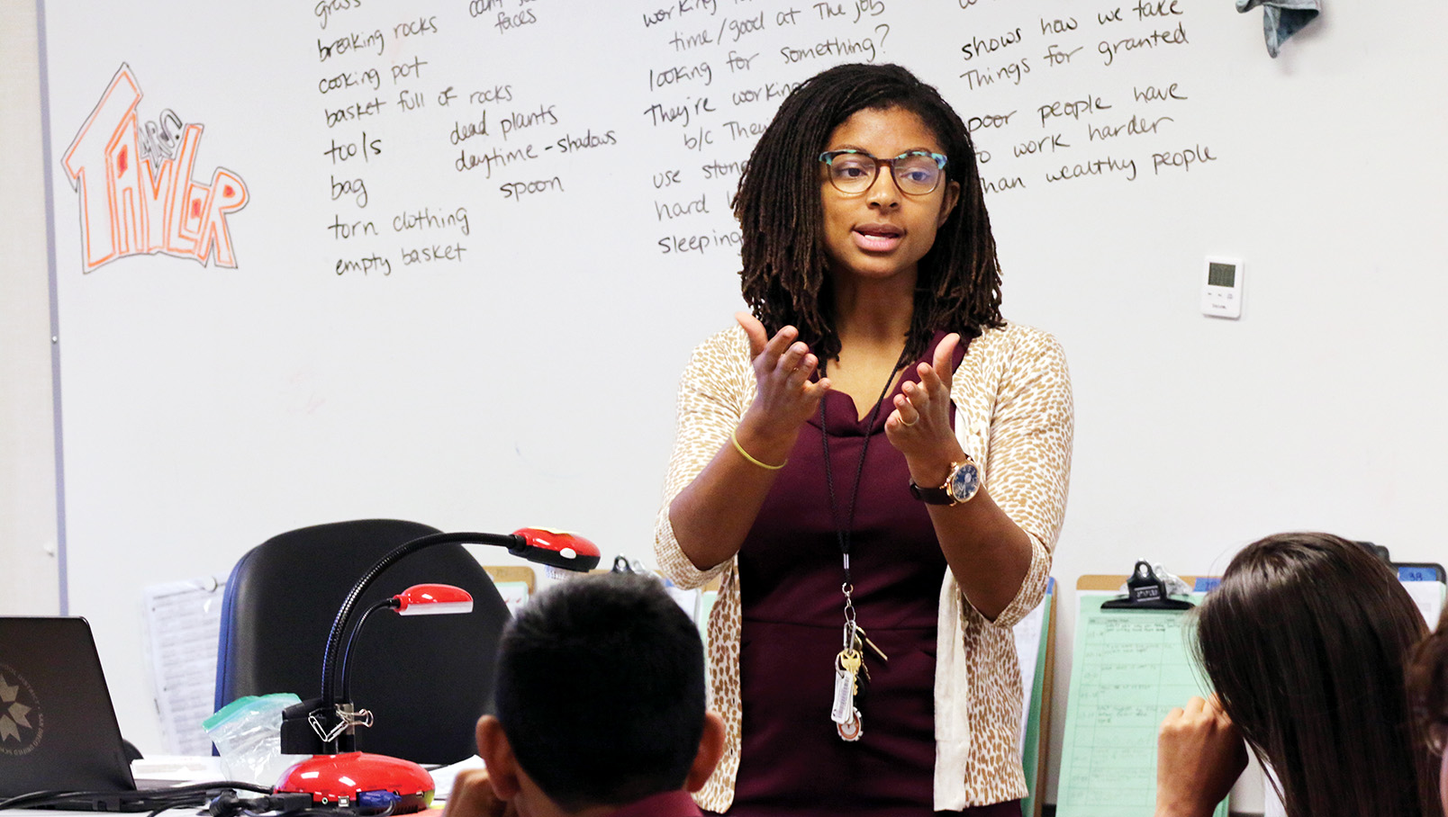 Person in a classroom setting giving a lecture, with a whiteboard full of notes in the background and students seated in front.