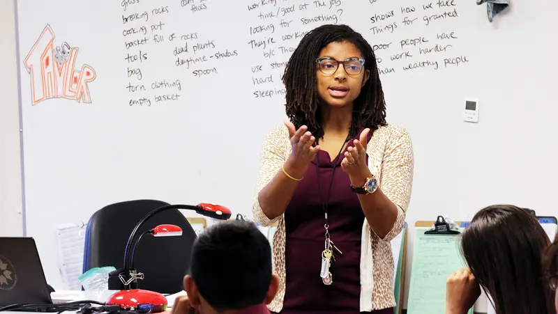 Person in a classroom setting giving a lecture, with a whiteboard full of notes in the background and students seated in front. Person in a classroom setting giving a lecture, with a whiteboard full of notes in the background and students seated in front.