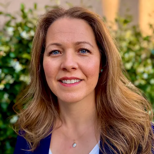 A person's headshot with a blurred face. They have long, wavy hair and are wearing a blue jacket with a white top and a necklace. The background consists of greenery with white flowers. A person's headshot with a blurred face. They have long, wavy hair and are wearing a blue jacket with a white top and a necklace. The background consists of greenery with white flowers.