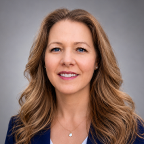 A person's headshot with a blurred face. They have long, wavy hair and are wearing a blue jacket with a white top and a necklace. The background consists of greenery with white flowers.