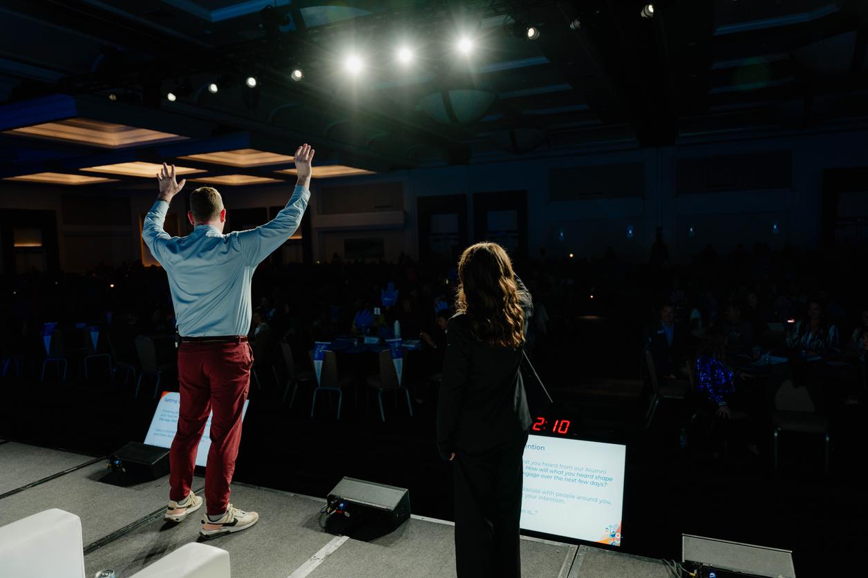 Two individuals standing on a stage facing an audience, under bright lights in a large room with multiple tables. Two individuals standing on a stage facing an audience, under bright lights in a large room with multiple tables.