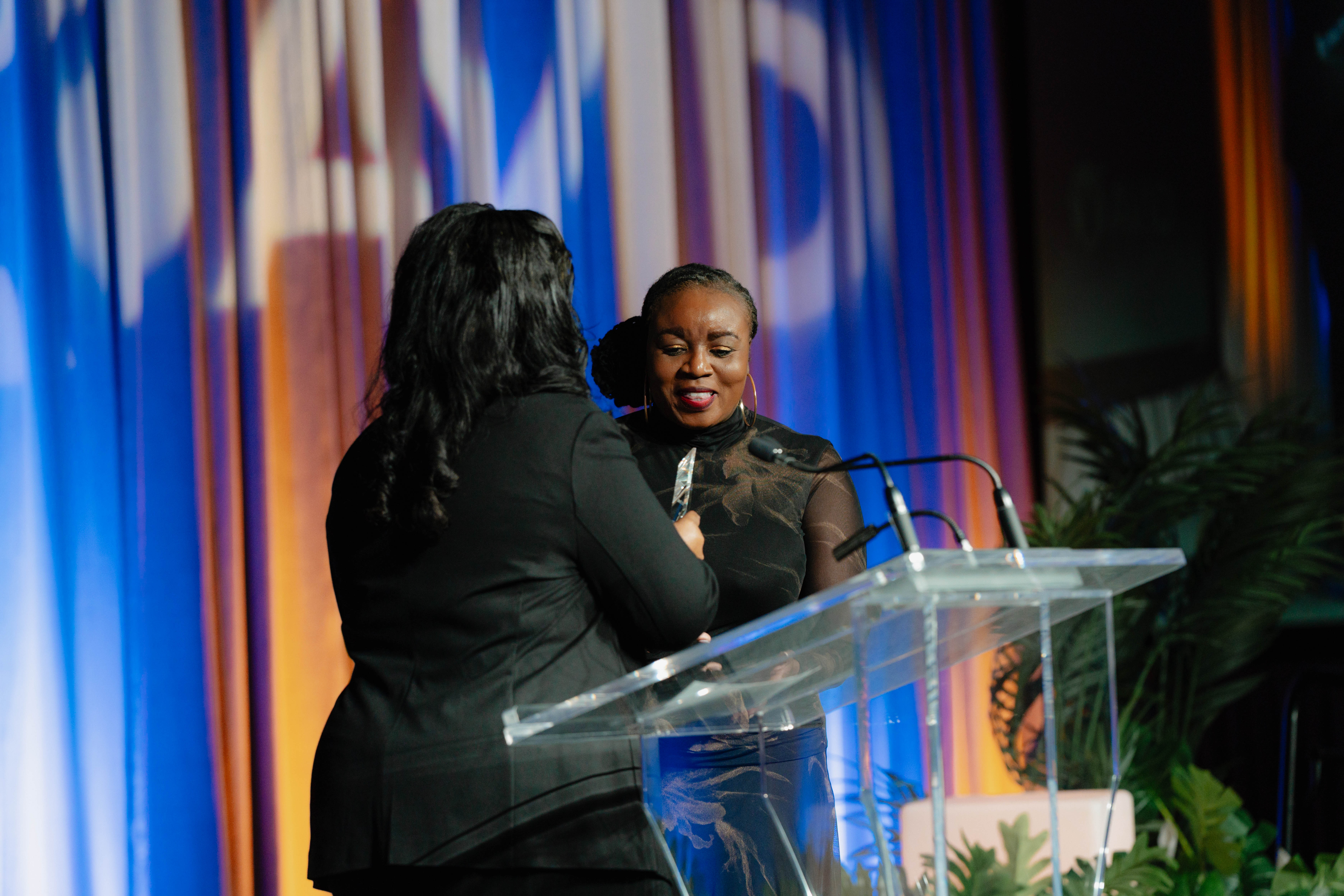 Two people standing at a podium with a microphone, one holding a glass award, with a colorful curtain backdrop behind them.