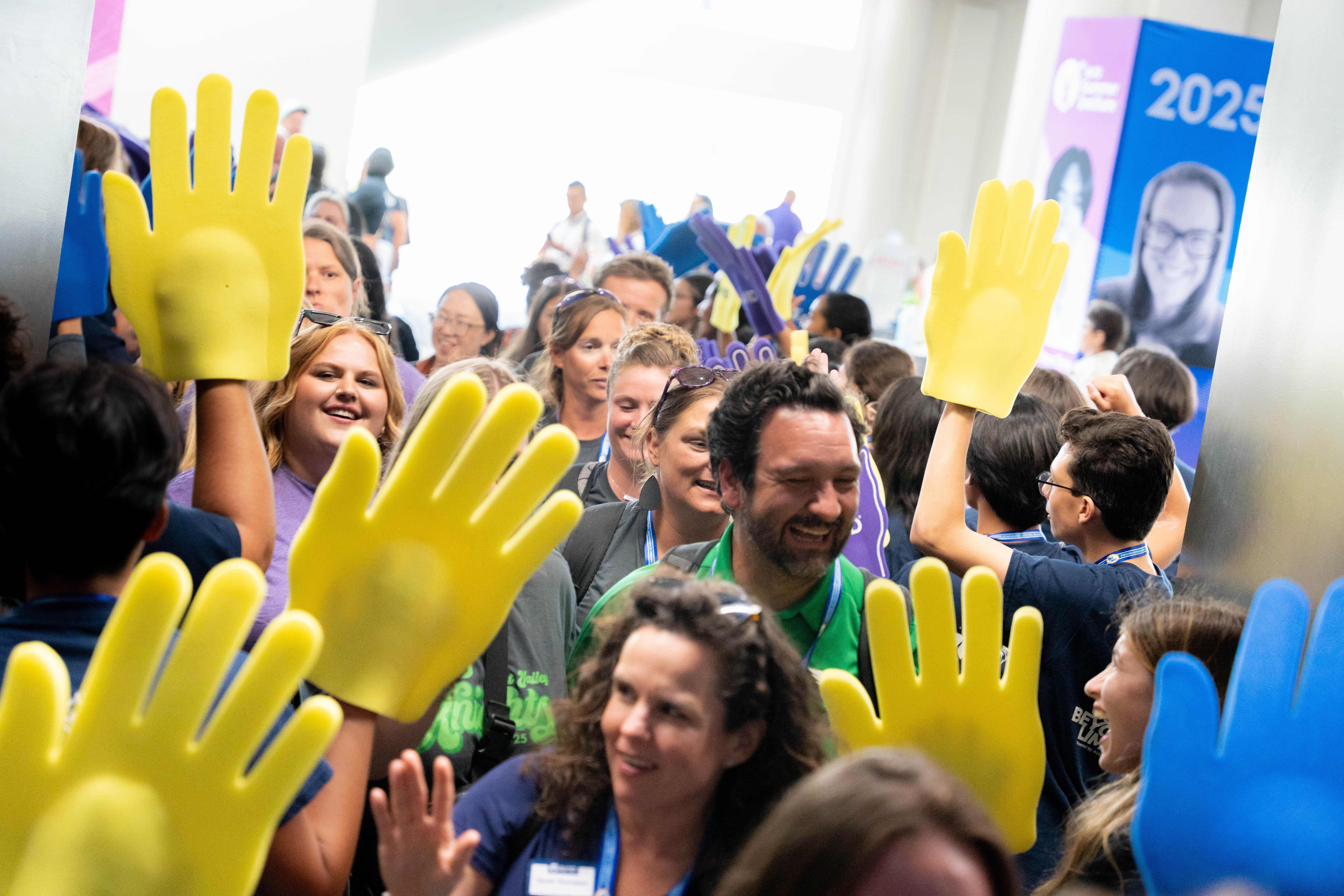 A crowded event with numerous people raising large yellow foam hands, standing in a brightly lit indoor setting.