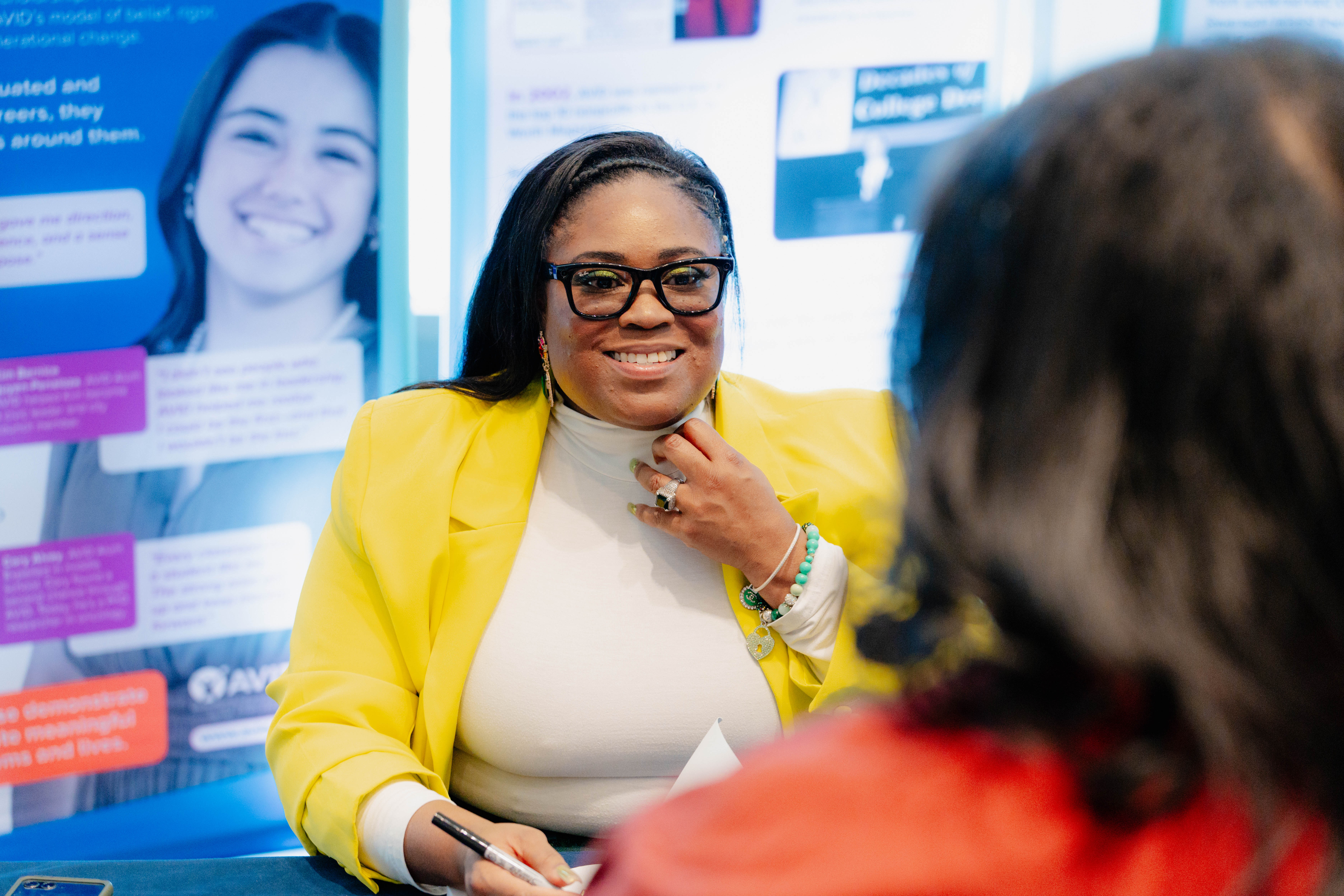Person in a yellow blazer sitting at a desk with informational posters in the background.