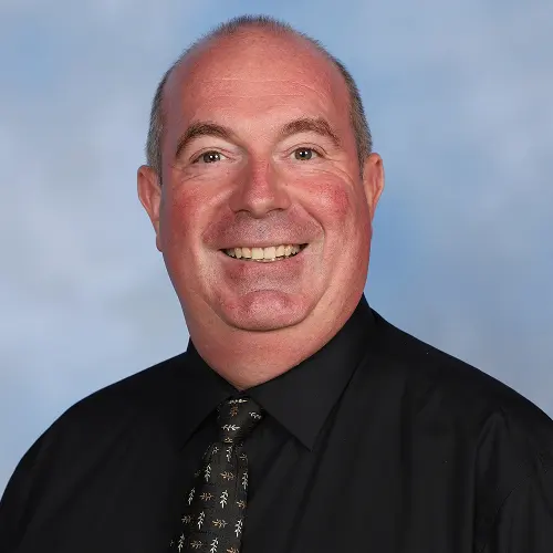 A blurred headshot of a person wearing a black shirt and patterned tie, against a blue-gray background. A blurred headshot of a person wearing a black shirt and patterned tie, against a blue-gray background.