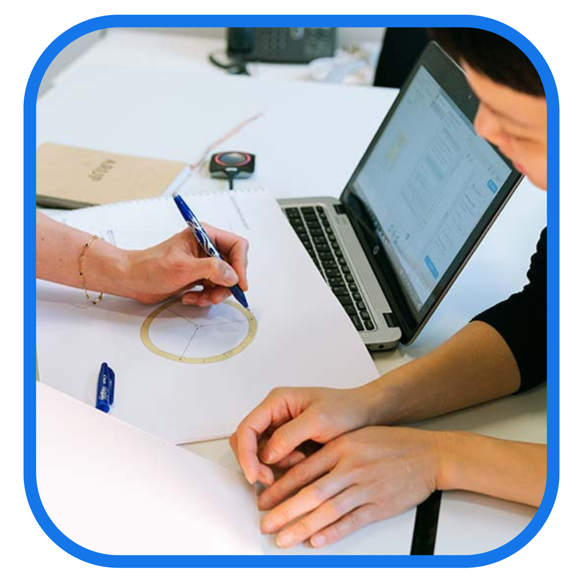 Two individuals collaborating at a desk, one writing with a pen on paper and the other working on a laptop.