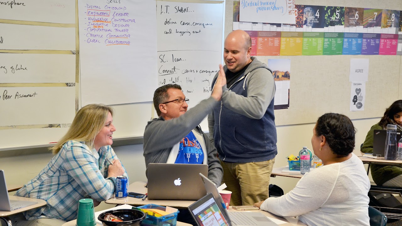 A group of people engaging in a collaborative activity within a classroom setting, featuring laptops and other materials on the desks.
