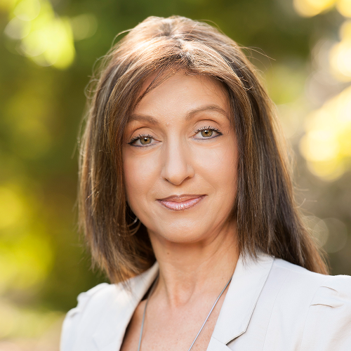 A person with shoulder-length hair wearing a light-colored outfit, standing outdoors with a blurred green background.