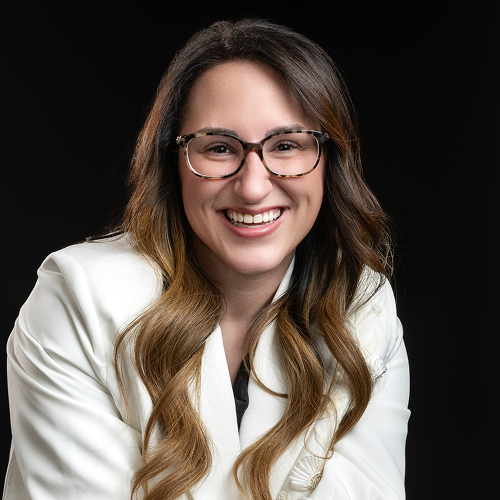 Individual with long, wavy hair wearing a white jacket against a black background, face blurred for anonymity.