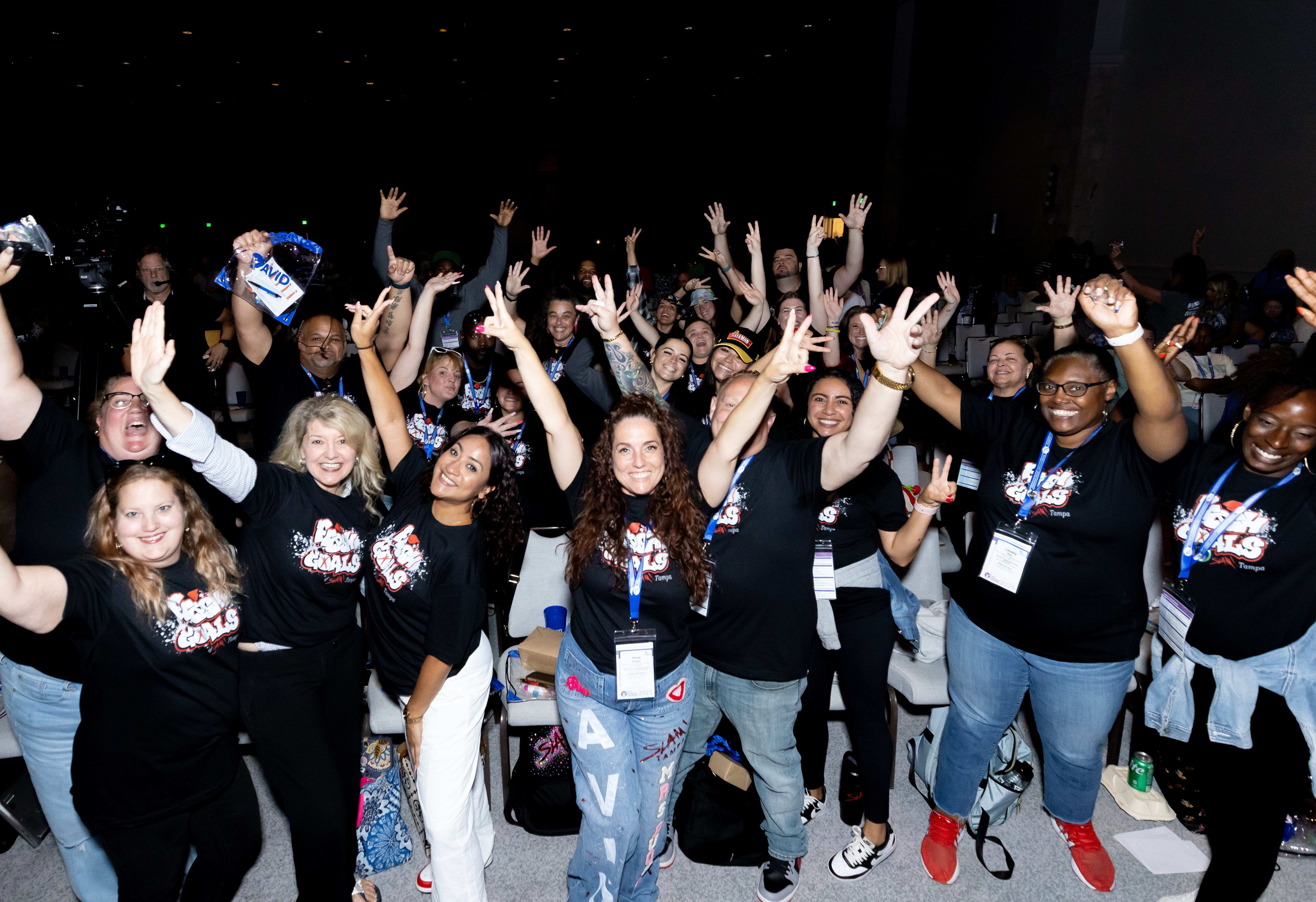 A large group of people wearing matching black shirts with graphic designs raise their hands in celebration at an indoor event.