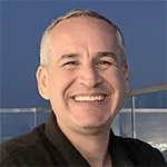Person with short hair wearing a dark colored shirt in front of a glass barrier with a blurred background.