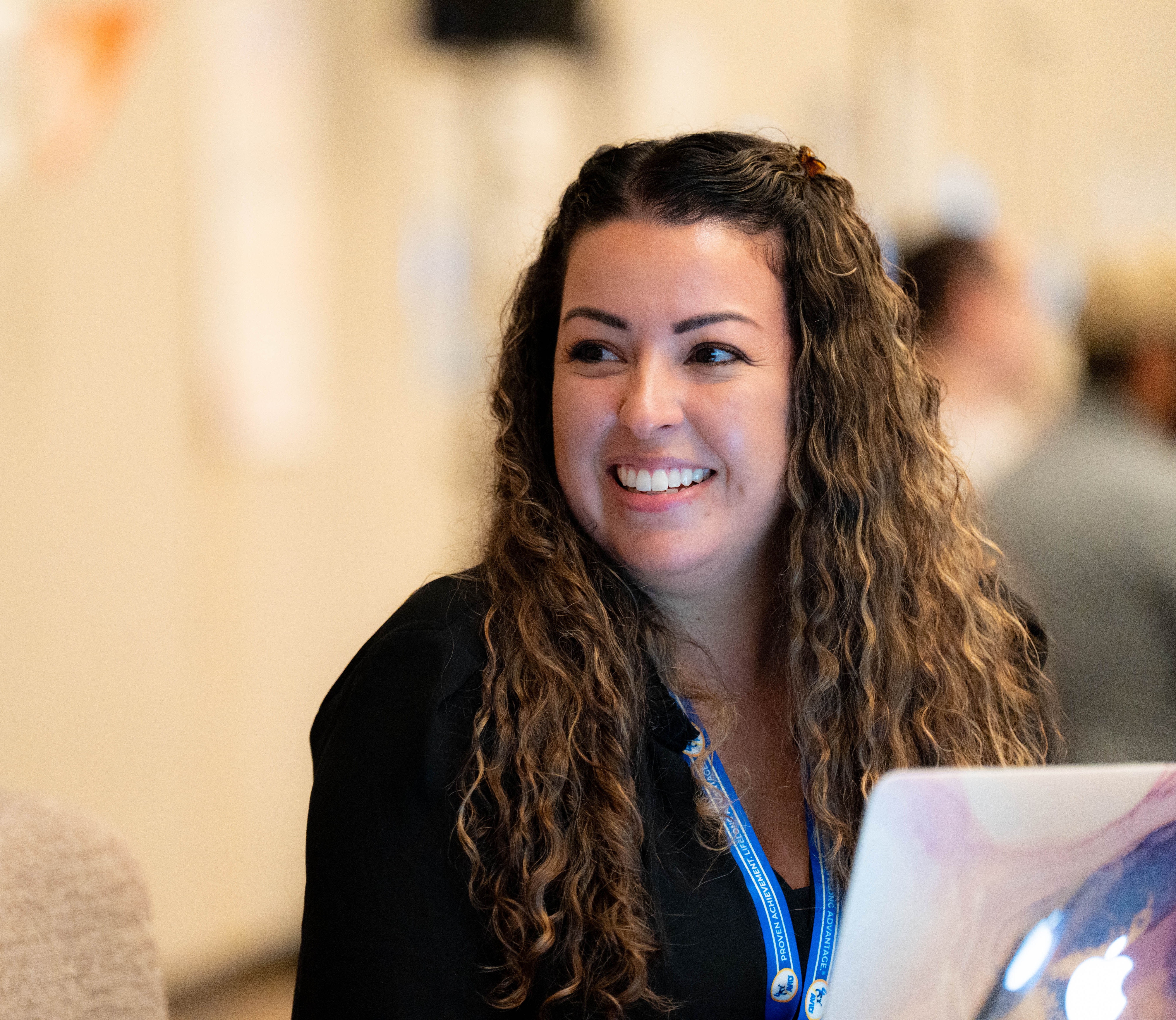 Person with long curly hair in a black outfit, wearing a lanyard with a visible badge, working on a laptop in a blurred indoor environment.