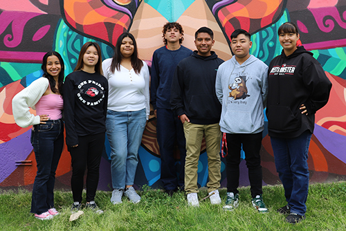 Group of seven people standing in front of a colorful mural.