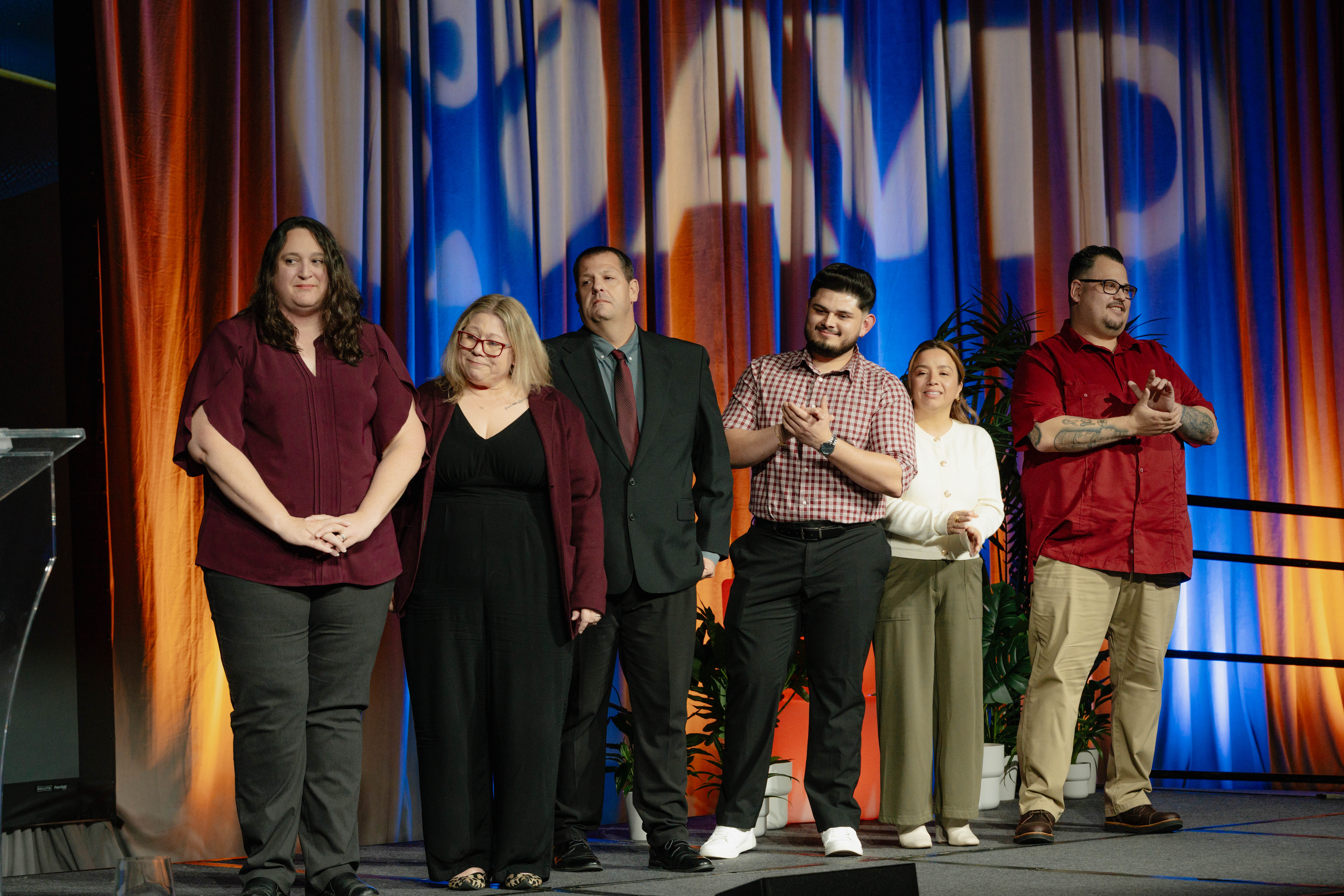 A group of six individuals standing on a stage in front of a colorful curtain backdrop, some clapping.