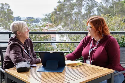 Two people sitting at an outdoor table, smiling and engaged in conversation, with a tablet and notebook on the table. Two people sitting at an outdoor table, smiling and engaged in conversation, with a tablet and notebook on the table.