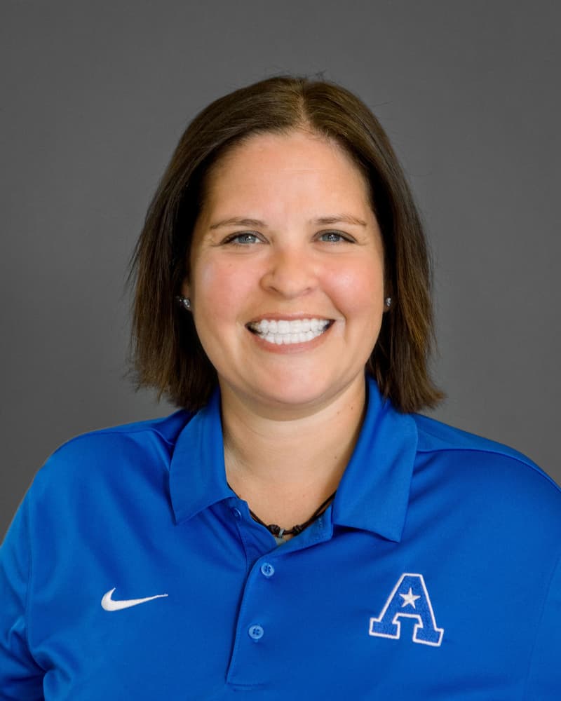 Person wearing a blue polo shirt with a logo, standing in front of a gray background. Person wearing a blue polo shirt with a logo, standing in front of a gray background.