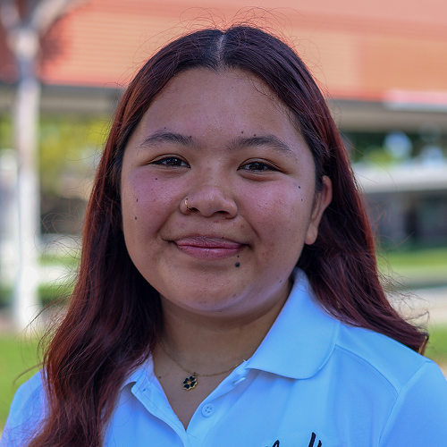 Individual with long hair wearing a white polo shirt, blurred face, outdoor background with building and trees.