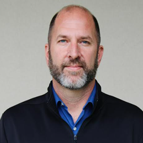 Person wearing a blue collar shirt and black jacket, standing against a neutral background. The face is blurred.