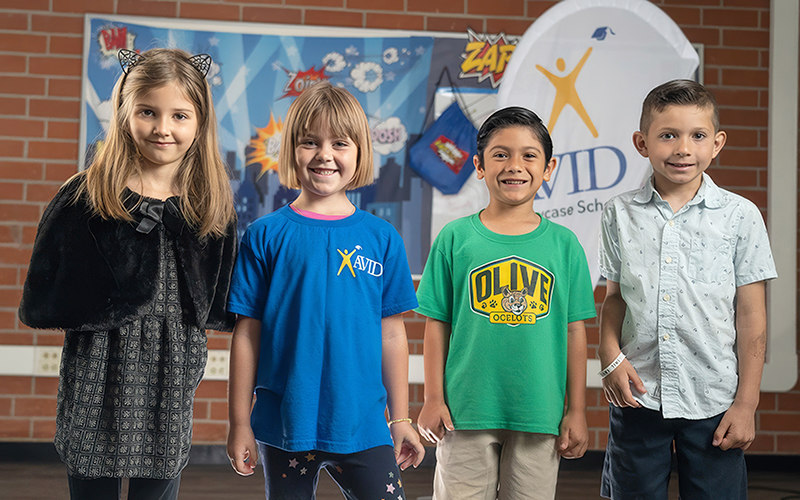 Group of four children standing, wearing casual clothing with a brick wall and educational posters in the background.