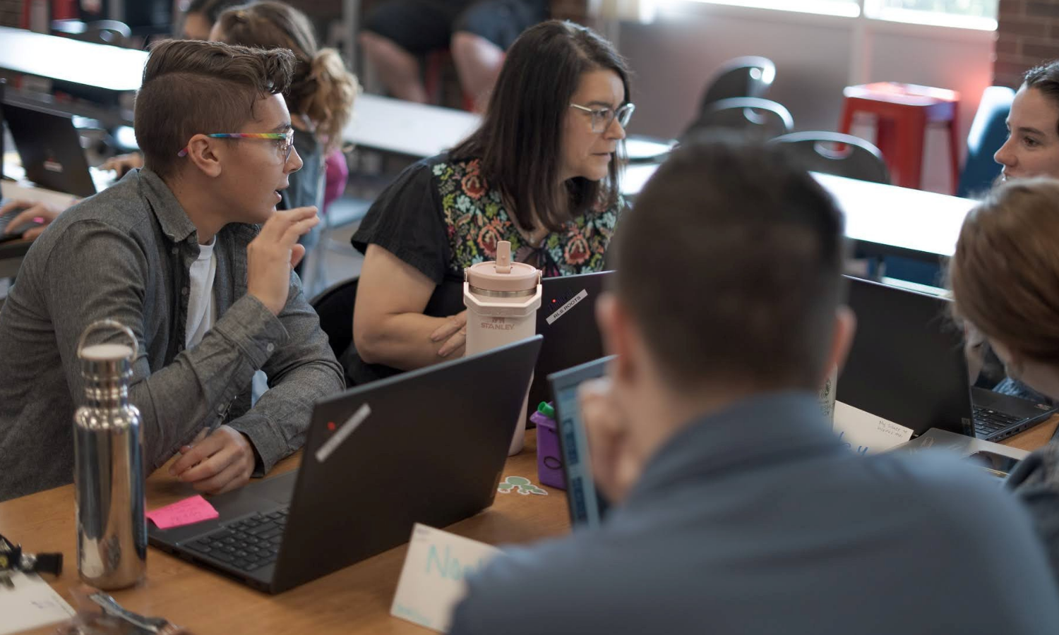 Group of people sitting at a table, using laptops and engaged in conversation in a classroom or office setting.