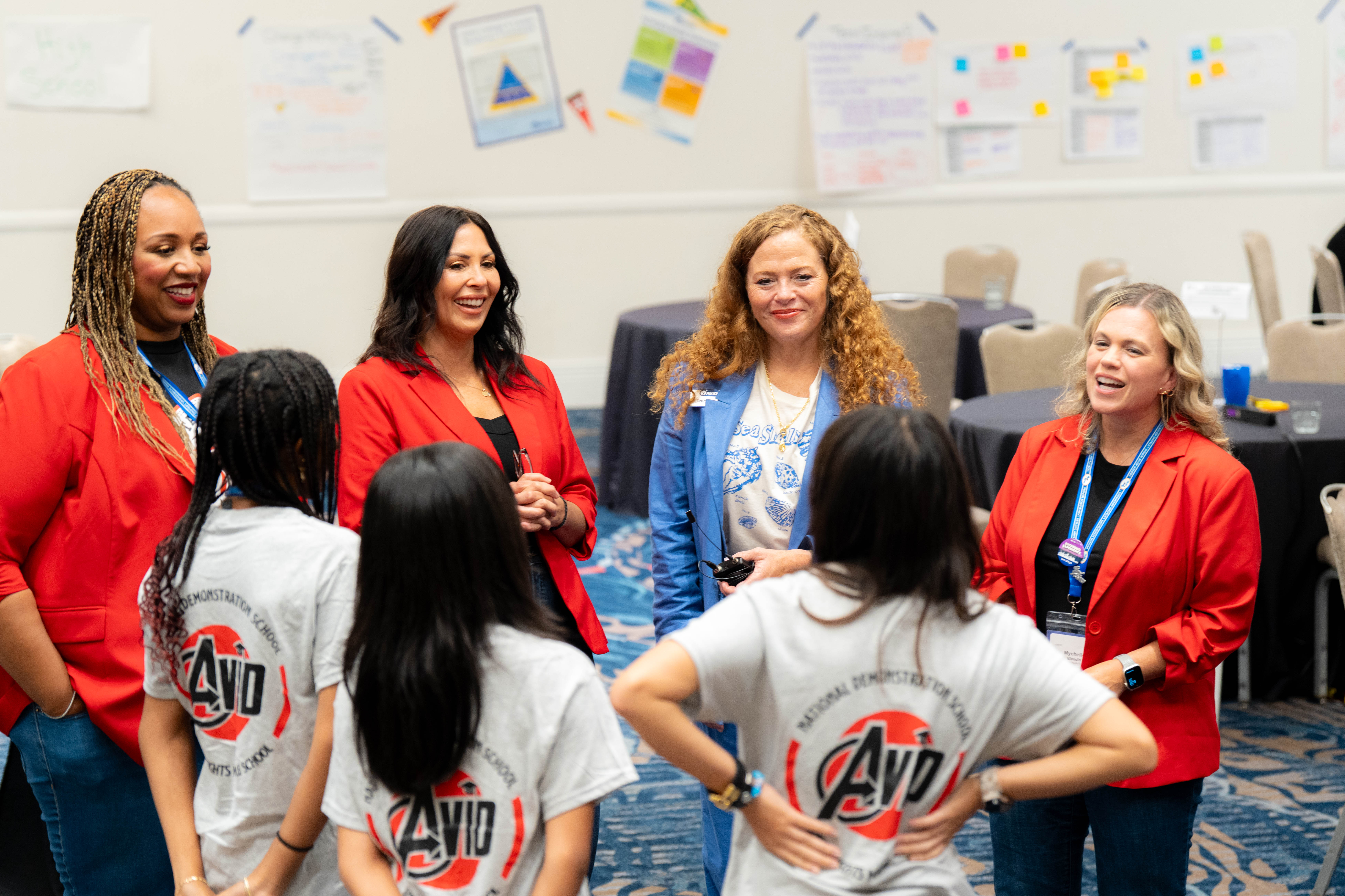 Several individuals in a conference setting, standing and talking. Some wear red jackets, others wear gray shirts with a logo.