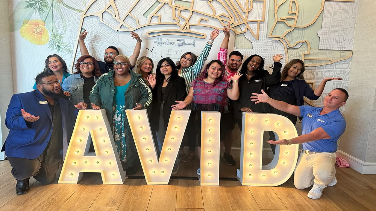 Group of people posing with illuminated letters spelling AVID against a decorative map background. Group of people posing with illuminated letters spelling AVID against a decorative map background.