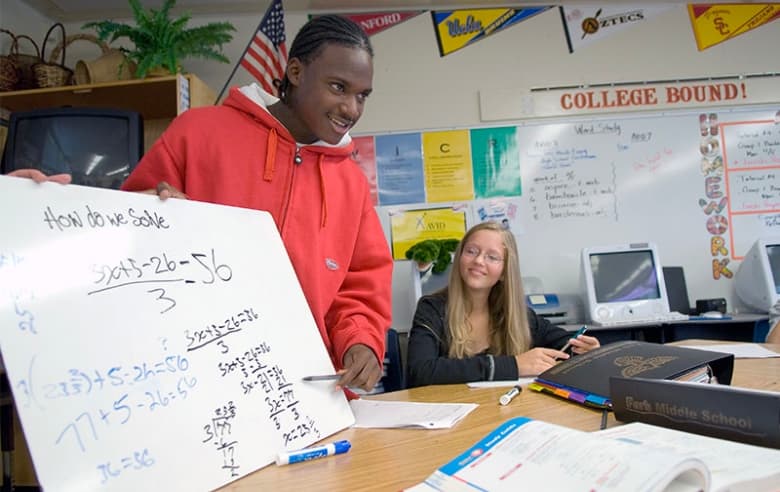 Two students in a classroom setting, one standing and presenting a whiteboard with math equations, the other sitting and writing at a desk, surrounded by school materials and a 'College Bound!' sign in the background. Two students in a classroom setting, one standing and presenting a whiteboard with math equations, the other sitting and writing at a desk, surrounded by school materials and a 'College Bound!' sign in the background.