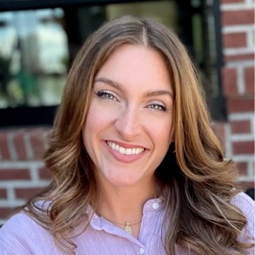 A person with long, wavy hair wearing a collared shirt against a background of brick and blurred elements.