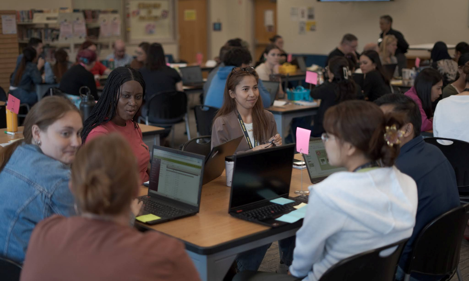 A group of people seated at tables in a classroom or meeting room, engaging in conversation and using laptops.